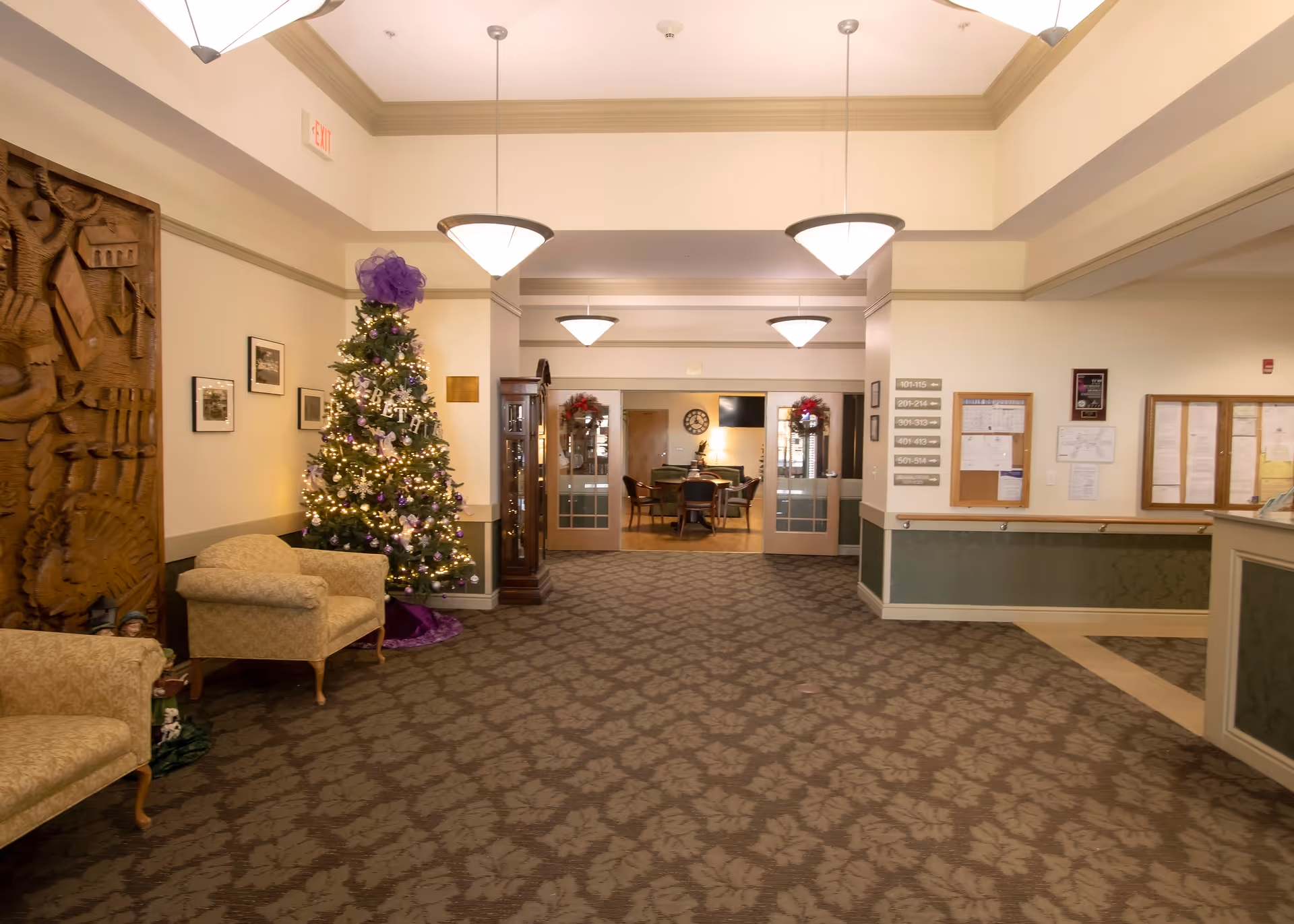 Interior view of a senior living facility lobby decorated for the holidays with a Christmas tree adorned with lights and purple decorations. The space features patterned carpet, beige armchairs, a wooden carved panel on the wall, and hanging pendant lights. In the background, there is a room with a round table and chairs, wall clock, and holiday wreaths on the glass doors.