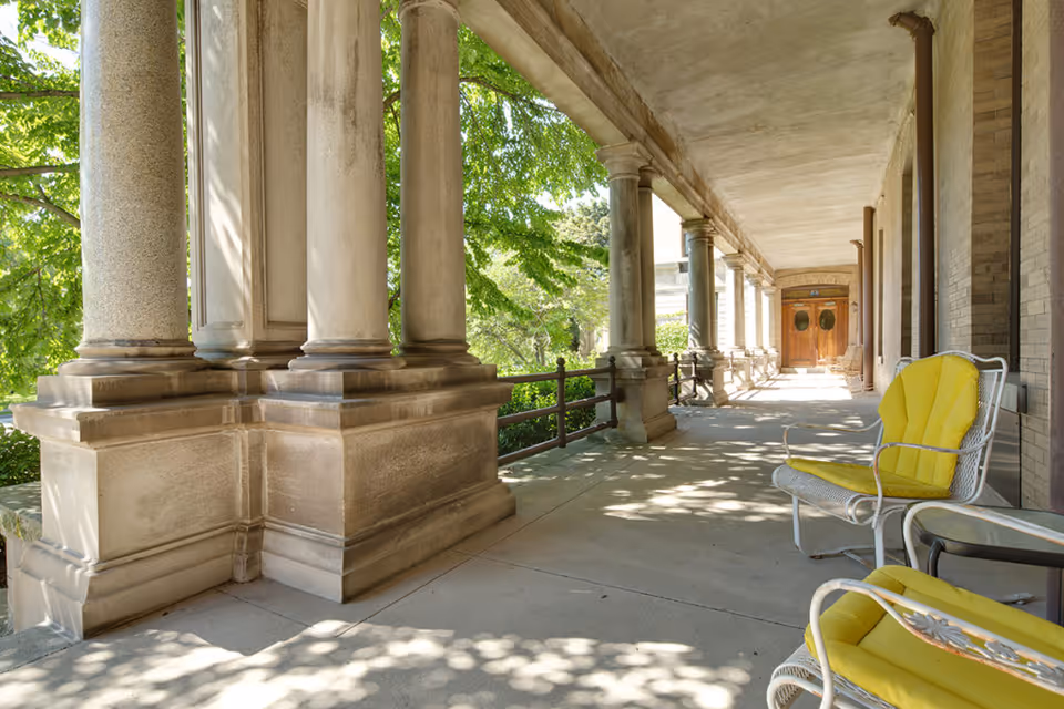 A shaded outdoor covered walkway with large stone columns and a concrete floor. Two white metal chairs with bright yellow cushions are positioned on the right side near a small table. Green trees and foliage are visible beyond the columns, and wooden double doors are seen at the far end of the walkway.