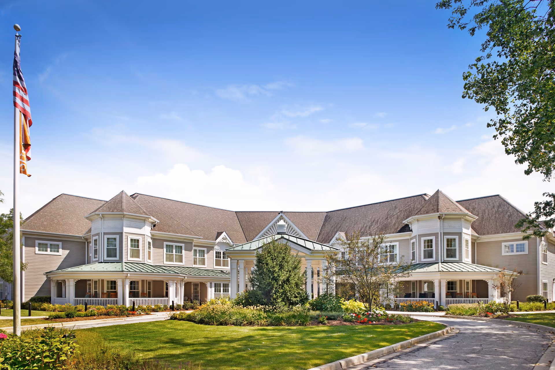 Front exterior view of Sunrise of Flossmoor, a large senior living facility with a well-maintained lawn, a circular driveway, and a flagpole with the American flag. The building features multiple windows, a peaked roof, and a covered entrance with white columns surrounded by greenery and flowers.
