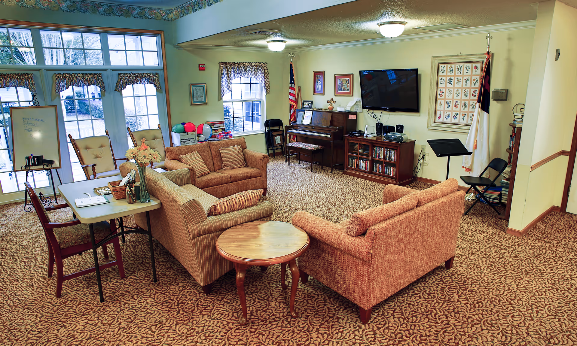 A cozy common area in a senior living facility with patterned carpet, two cushioned sofas, a small round wooden table, and a rectangular table with chairs. There is a piano against the wall with framed pictures above it, a flat-screen TV mounted on the wall, and an American flag in the corner. Large windows and glass doors let in natural light, and a whiteboard with some writing is visible near the doors.