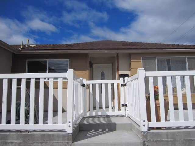 Front view of a single-story residential building with a white gate and railing leading to a white front door. The building has beige and light brown exterior walls, two windows on either side of the door, and a clear blue sky with some clouds above.
