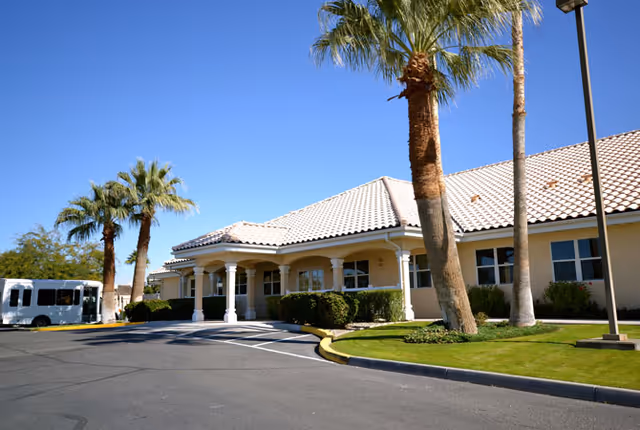Exterior view of Avista Senior Living Yuma building with a tiled roof, palm trees, and a driveway under a clear blue sky.