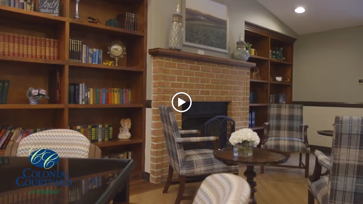 A cozy interior lounge area featuring a brick fireplace flanked by wooden bookshelves filled with books and decorative items. There are several plaid upholstered armchairs arranged around a small wooden table with a vase of white flowers. The room has warm lighting and a comfortable, inviting atmosphere.