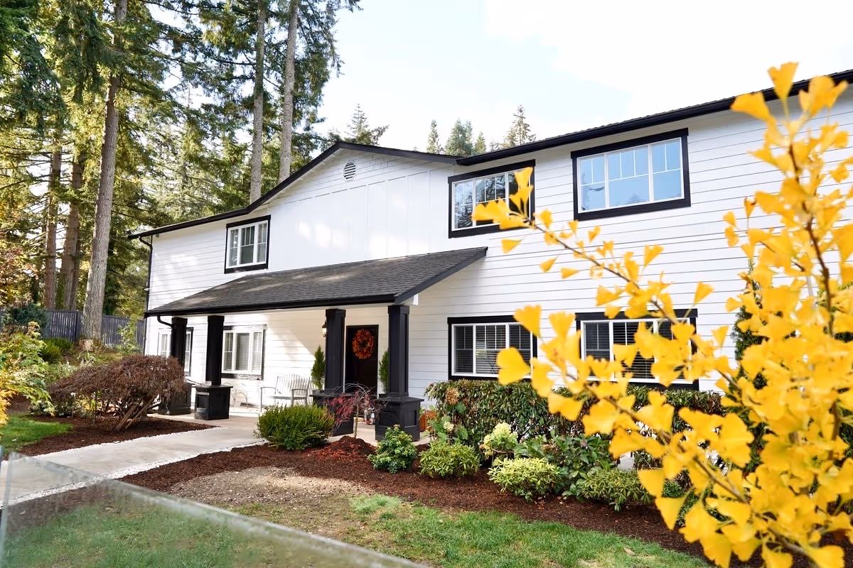 Exterior view of a two-story white senior living facility with black trim around the windows and doors. The building has a covered porch with black pillars and outdoor seating. There are landscaped bushes and plants in front of the building, with tall trees in the background and a yellow-leaved plant in the foreground.