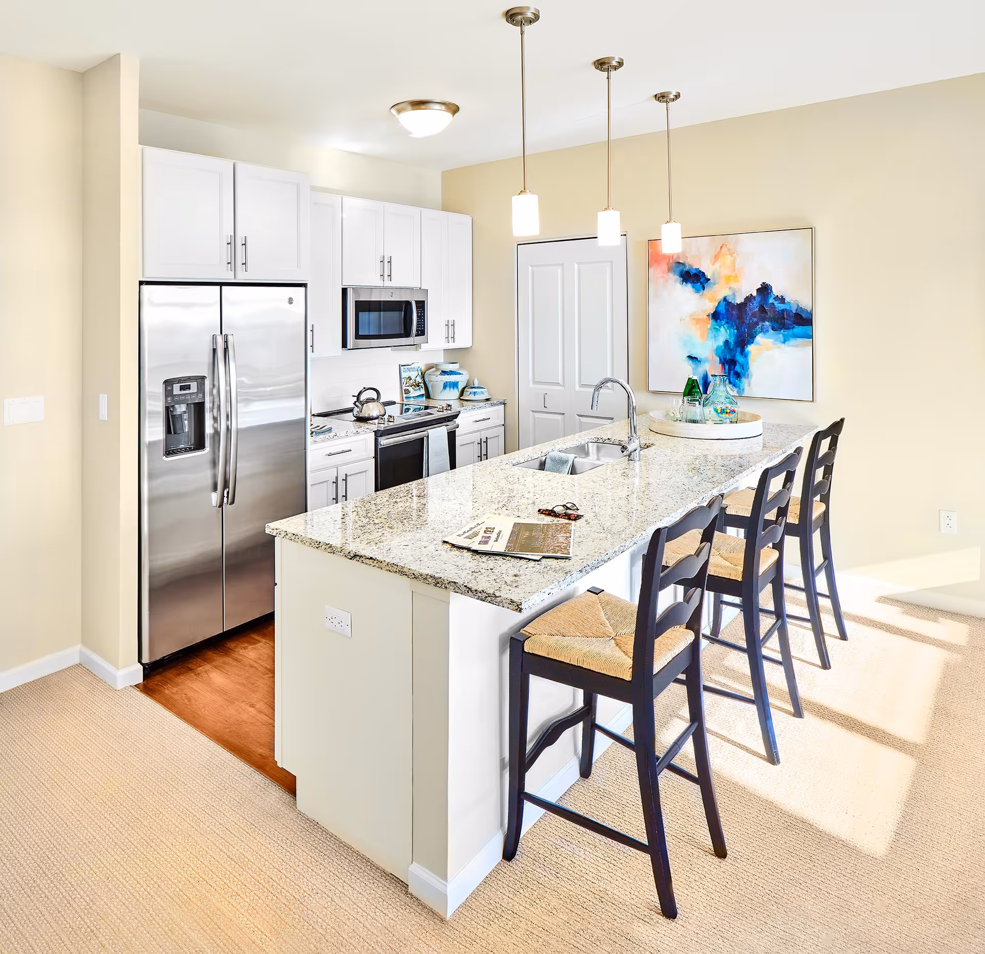 Bright modern kitchen with white cabinets, stainless steel refrigerator and microwave, granite countertop island with sink, three black bar stools with woven seats, and abstract blue and orange painting on the wall.