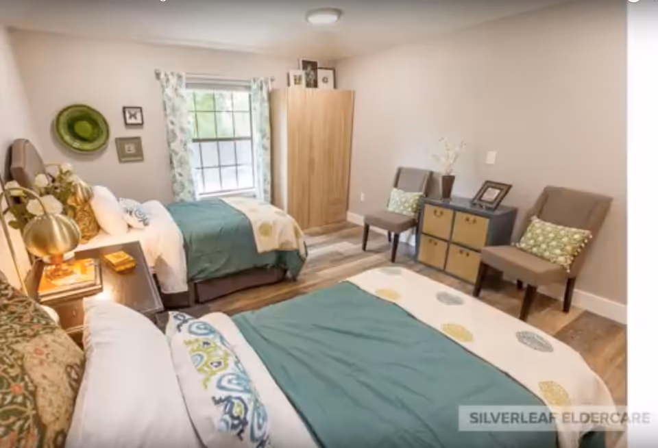 A bright bedroom with two twin beds, each with teal and patterned bedding. There is a window with floral curtains between the beds, a wooden wardrobe in the corner, two chairs with green patterned pillows, and a small cabinet with decorative items on top. The room has light-colored walls and wooden flooring.
