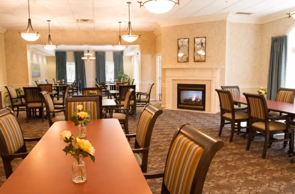 A spacious dining room with multiple wooden tables and striped cushioned chairs. Each table has a small vase with yellow flowers. The room features a carpeted floor, beige patterned walls, large windows with blue curtains, and a fireplace with a fire burning. Ceiling lights illuminate the room warmly.