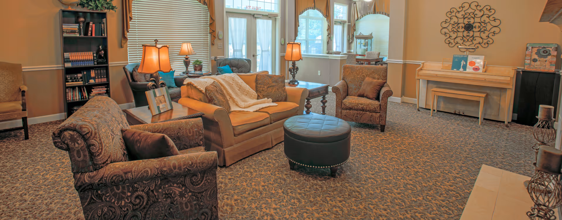 A cozy living room with patterned carpet, a sofa with a throw blanket, two armchairs, a round ottoman, side tables with lamps, a bookshelf, a piano, and large windows letting in natural light.