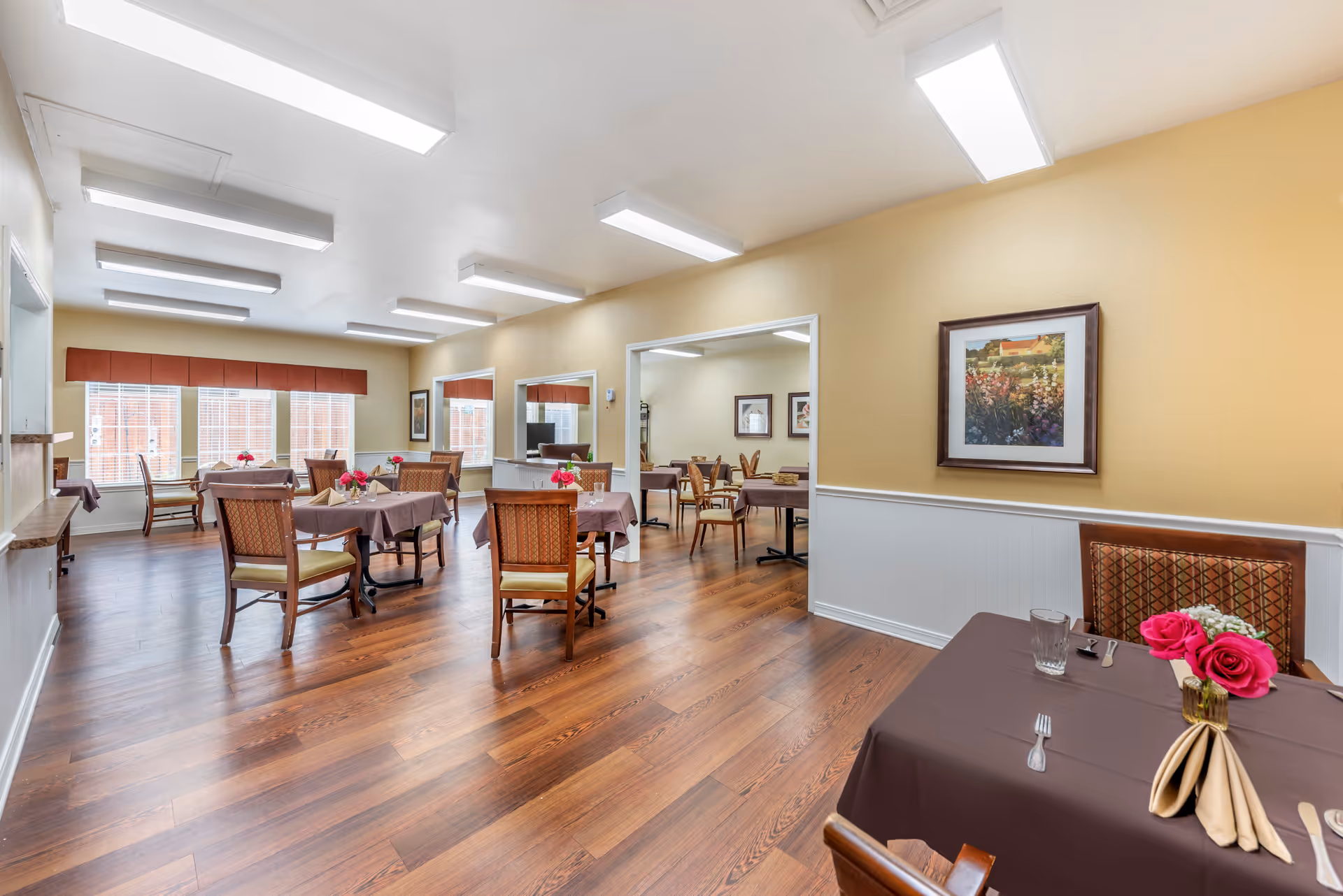 A bright dining room with several tables covered in brown tablecloths, each set with napkins, utensils, glasses, and small floral centerpieces. The room has wooden flooring, beige walls with white wainscoting, and large windows with red valances allowing natural light to enter. There are framed paintings on the walls and fluorescent ceiling lights.
