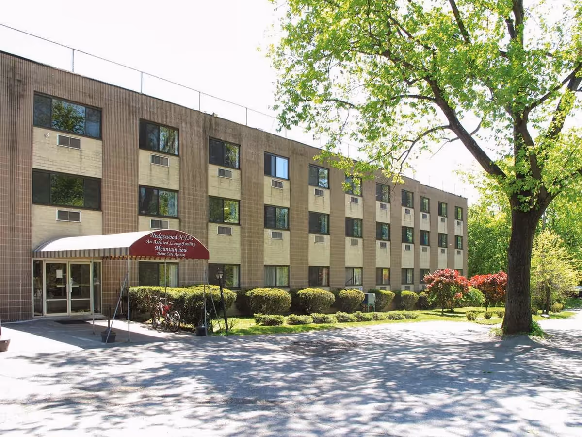Exterior view of a three-story assisted living facility building with multiple windows, a red canopy entrance labeled Hedgewood HFA, surrounded by green trees and bushes on a sunny day.