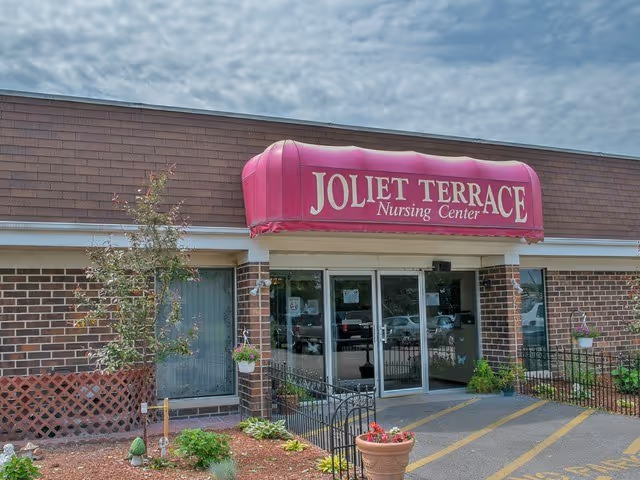 Entrance of Joliet Terrace Nursing Center with a red awning displaying the facility name above glass double doors. The building has a brick exterior with some small plants and flowers near the entrance.