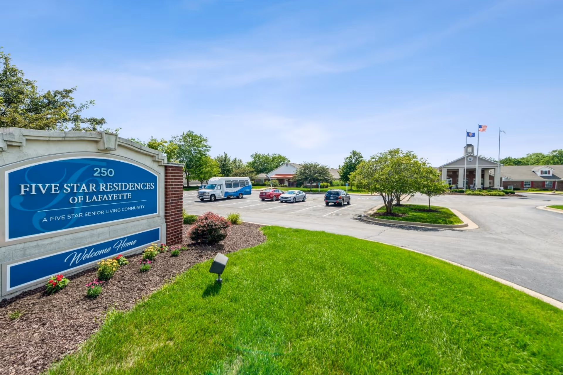 Exterior view of Five Star Residences of Lafayette senior living community showing the entrance sign, parking lot with several vehicles, green lawn, trees, and the main building with flagpoles displaying flags under a clear blue sky.