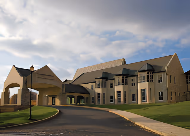 Exterior view of a large building with a covered entrance driveway, surrounded by green grass under a partly cloudy sky.
