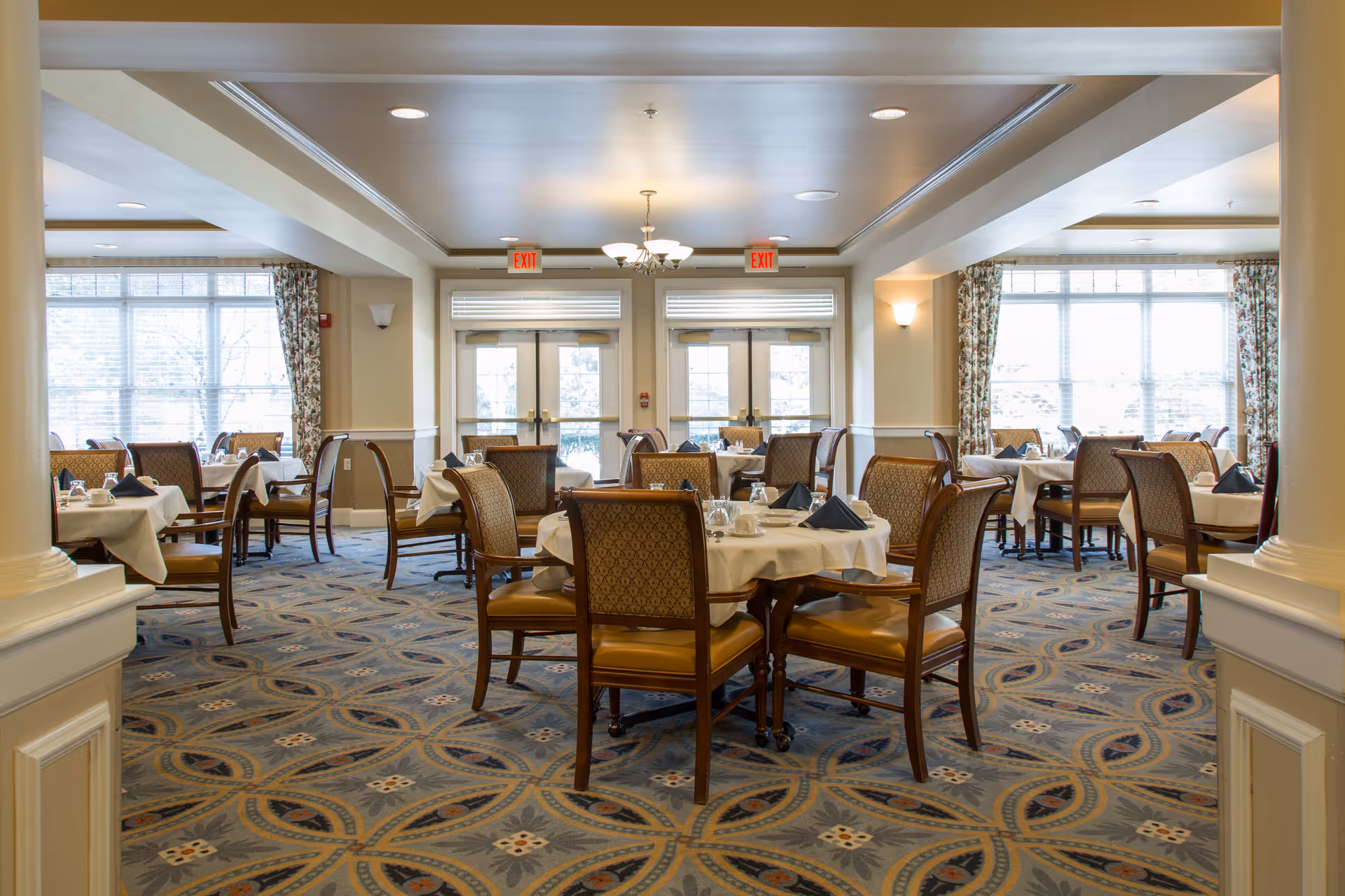 A spacious dining room with multiple round tables covered with white tablecloths, each set with napkins, cups, and utensils. The room has large windows with floral curtains allowing natural light to fill the space. The carpet has a decorative pattern, and there are exit signs above double doors at the far end of the room.