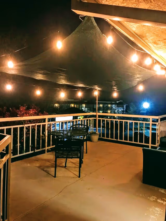 Outdoor patio area at night with string lights hanging above, a small table with two chairs, a potted plant, and a railing overlooking a parking lot and buildings in the background.