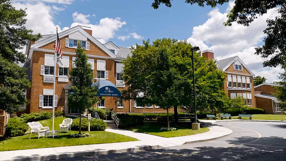 Exterior view of a large brick senior living facility building with white window shutters and a blue awning that reads 'Atria Welcome Home'. The building is surrounded by green trees, a well-maintained lawn, white outdoor chairs, and an American flag on a flagpole. The sky is partly cloudy with patches of blue.