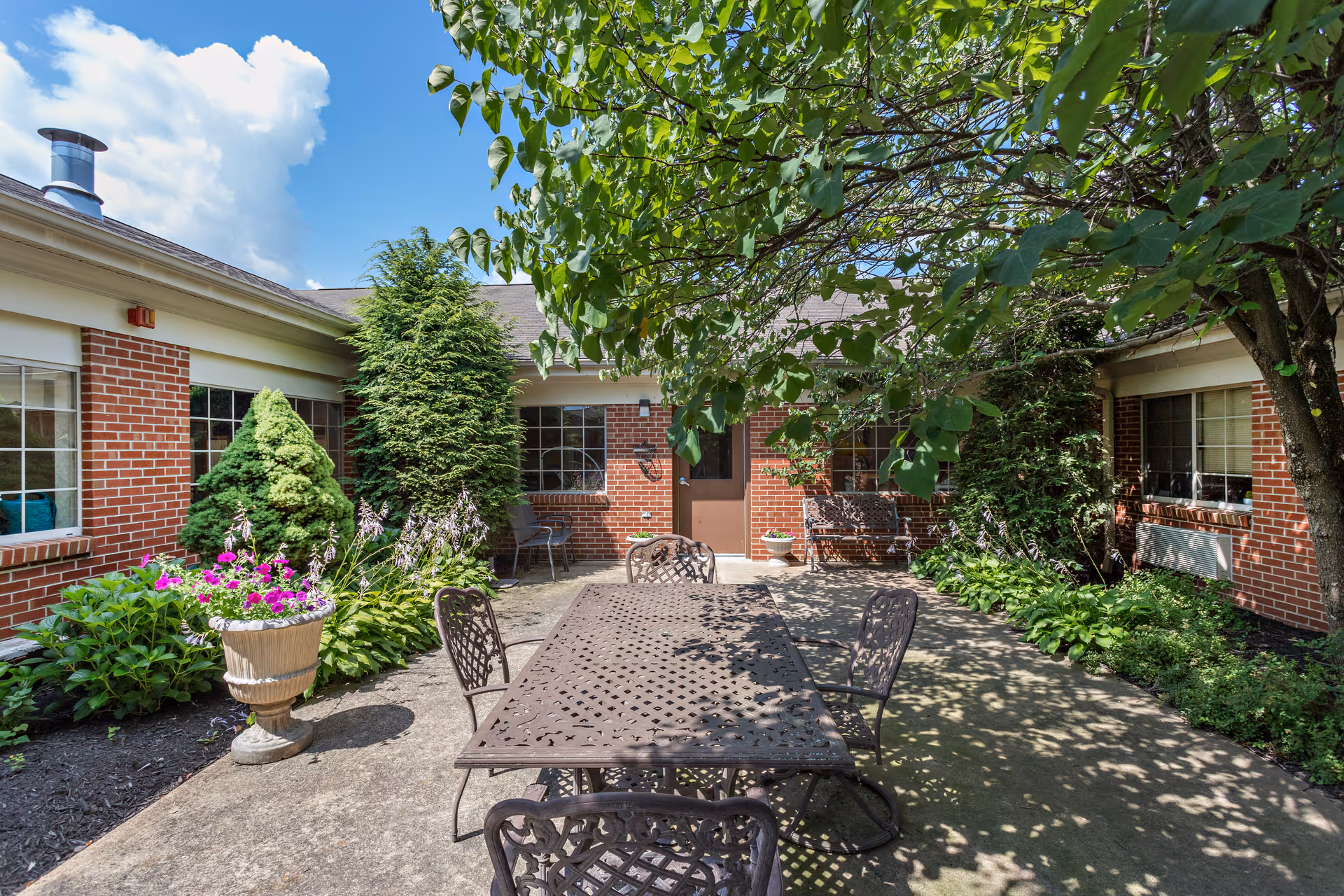 Outdoor patio area at North Strabane Rehabilitation & Wellness Center featuring a metal table with six matching chairs, surrounded by lush green plants and trees. The patio is bordered by a red brick building with multiple windows and a door in the center. A large tree provides shade over part of the seating area.