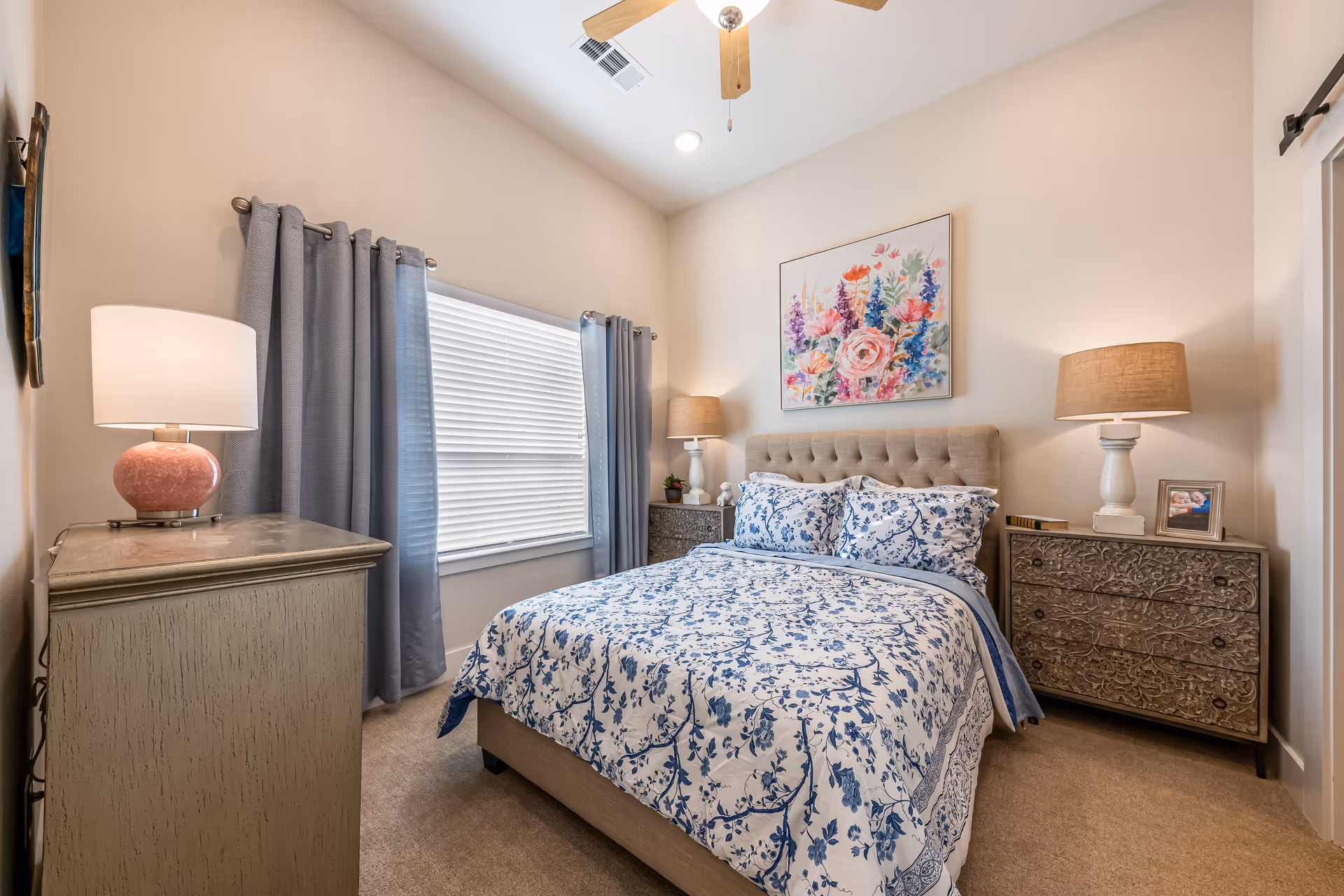 A cozy bedroom with a bed dressed in blue and white floral bedding, two matching nightstands with lamps on each side, a dresser with a pink lamp, a window with gray curtains, a ceiling fan, and a colorful floral painting on the wall above the bed.