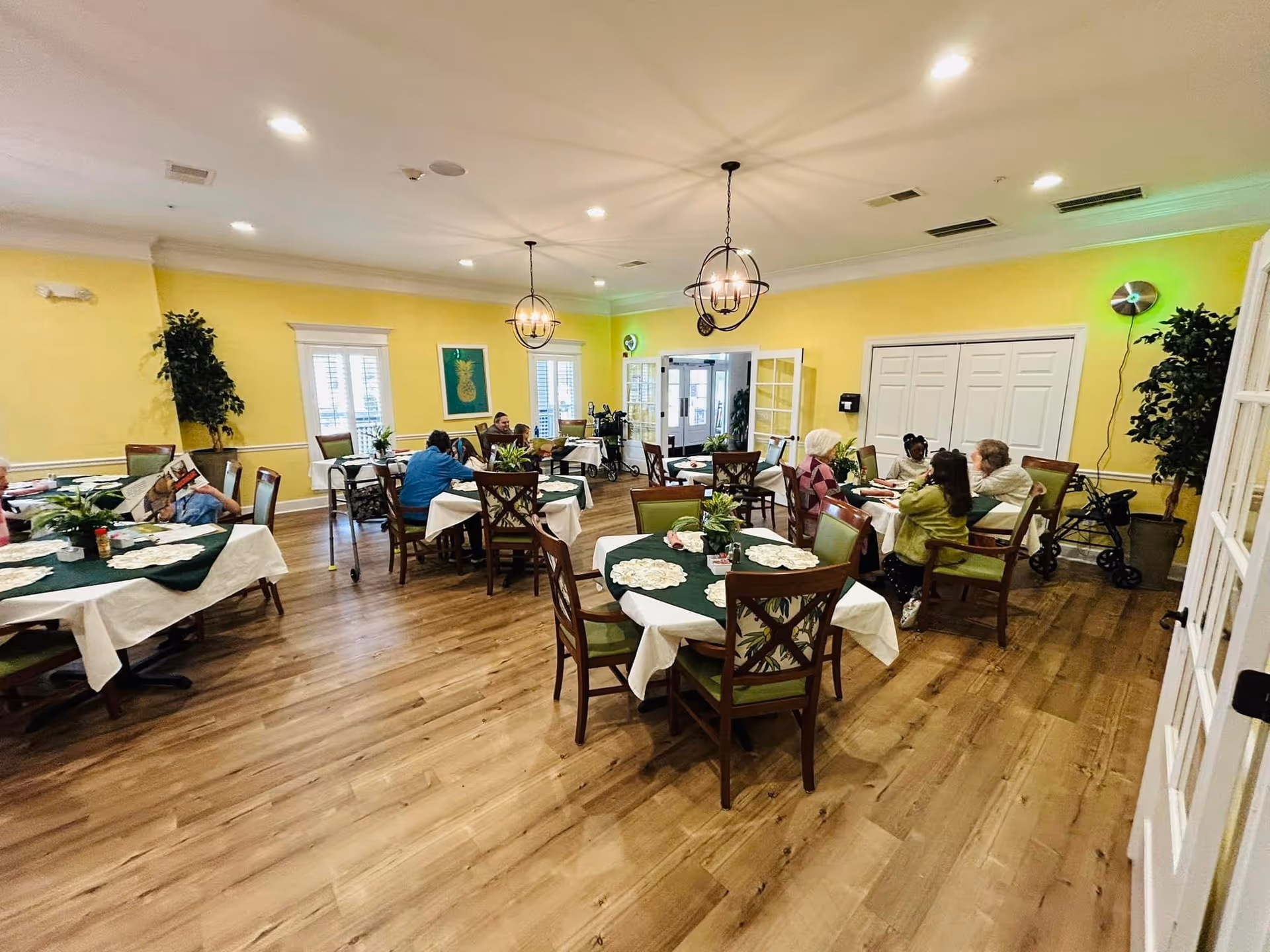 A bright dining room with yellow walls and wooden flooring. Several round tables are covered with white tablecloths and green table runners, each decorated with plants and doilies. Elderly residents are seated around the tables, some engaged in conversation and others reading. The room is well-lit with ceiling lights and two hanging chandeliers. There are large windows with white shutters and double doors leading outside.