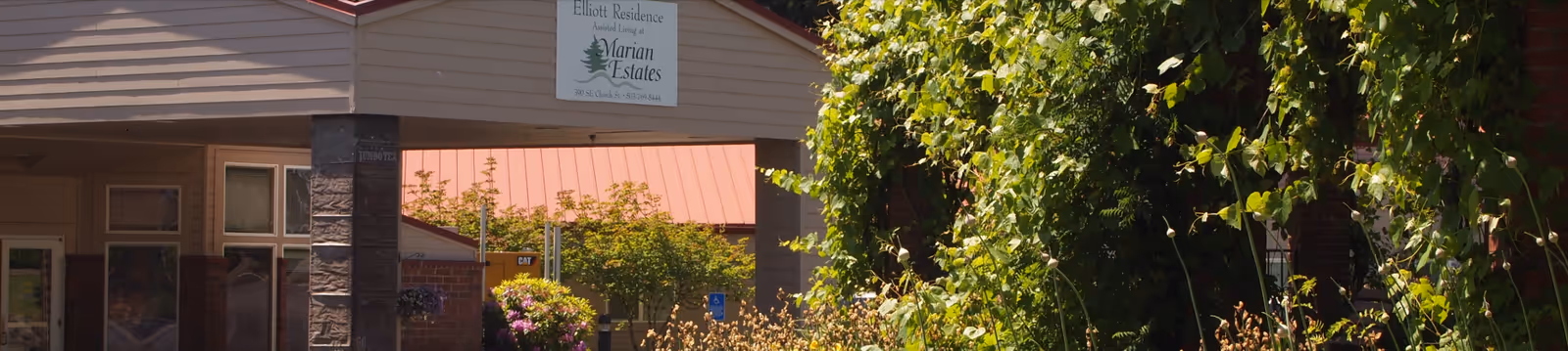 Exterior view of Marian Estates facility showing part of the building with a sign that reads 'Elliott Residence Marian Estates' surrounded by greenery and trees.