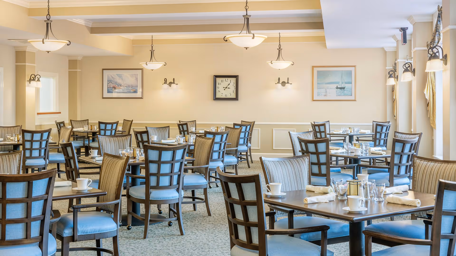 A bright and spacious dining room with multiple wooden tables and chairs featuring blue cushions. Each table is set with white cups, napkins, and silverware. The walls are painted a soft beige and decorated with framed paintings and a clock. Ceiling lights and wall sconces provide warm lighting.