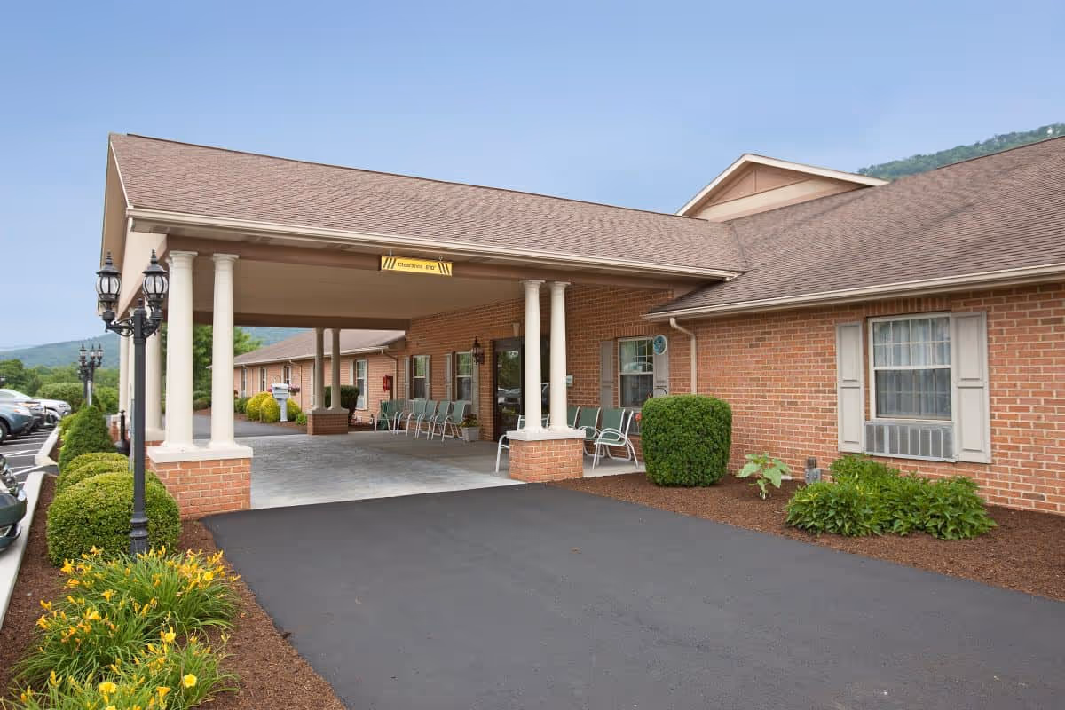 Exterior view of Colonial Courtyard at Bedford showing a covered entrance with white columns, brick walls, green chairs lined up along the wall, neatly trimmed bushes, yellow flowers, and a paved driveway under a clear sky.