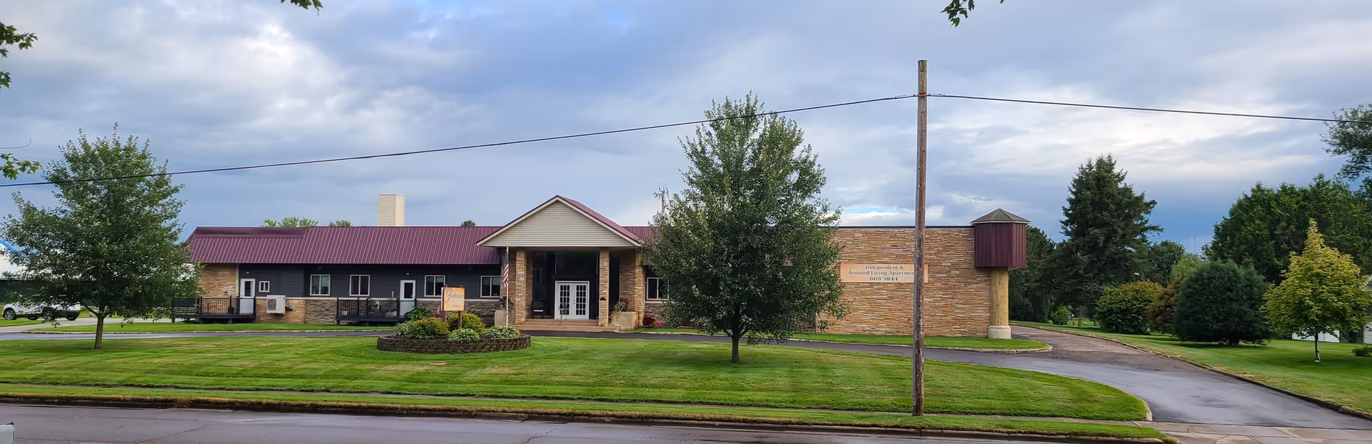 Front exterior view of Traditions Assisted Living building with a maroon roof, beige brick walls, a covered entrance with columns, surrounded by green grass, trees, and a cloudy sky.