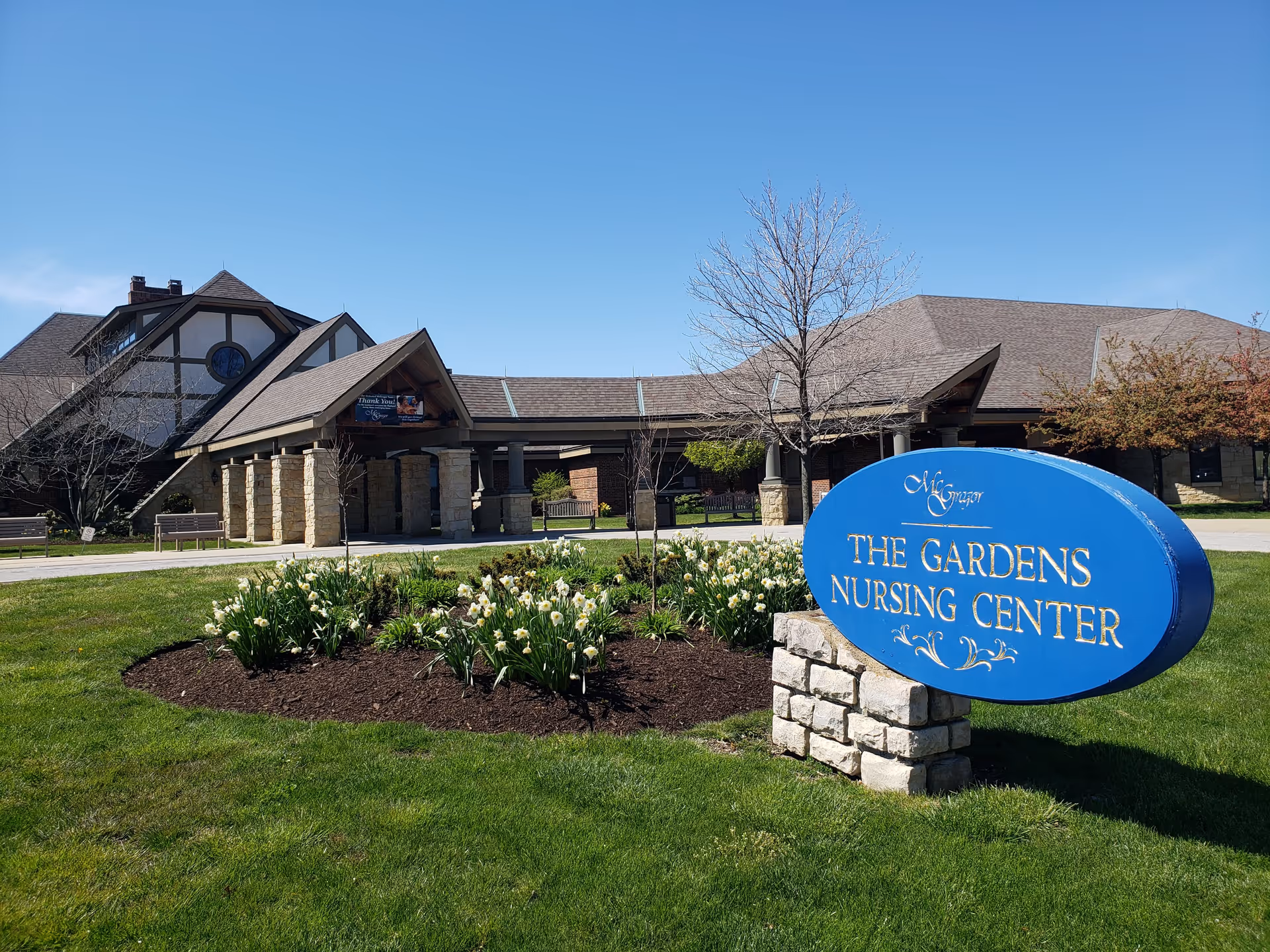 Exterior view of The Gardens Nursing Center at McGregor Assisted Living, featuring a blue sign with the facility's name in front of a landscaped flower bed with white flowers and a large building with a covered entrance in the background under a clear blue sky.