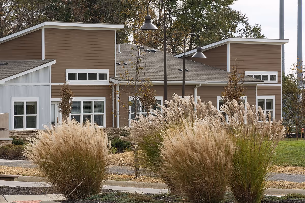 Exterior view of a modern senior living facility building with brown and white siding, multiple windows, and decorative tall grasses in the landscaped front area.