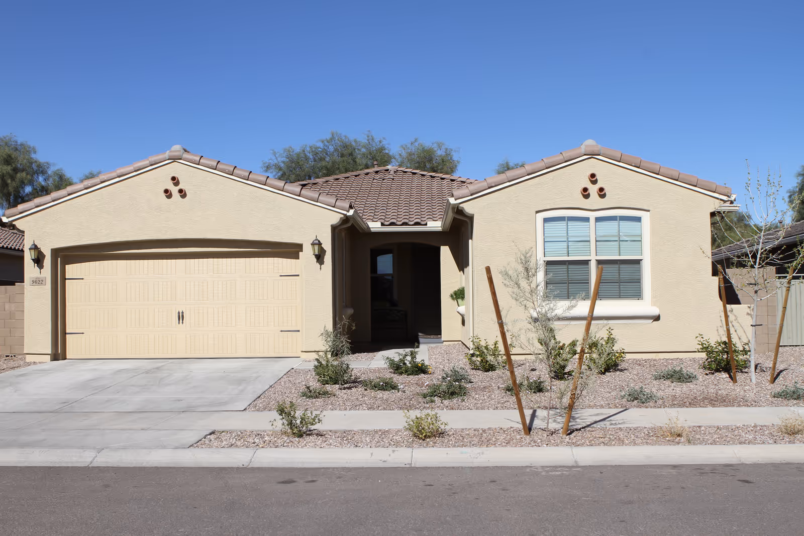 Front exterior view of a single-story beige house with a tiled roof, a two-car garage, a small front porch, and desert landscaping with small plants and gravel.