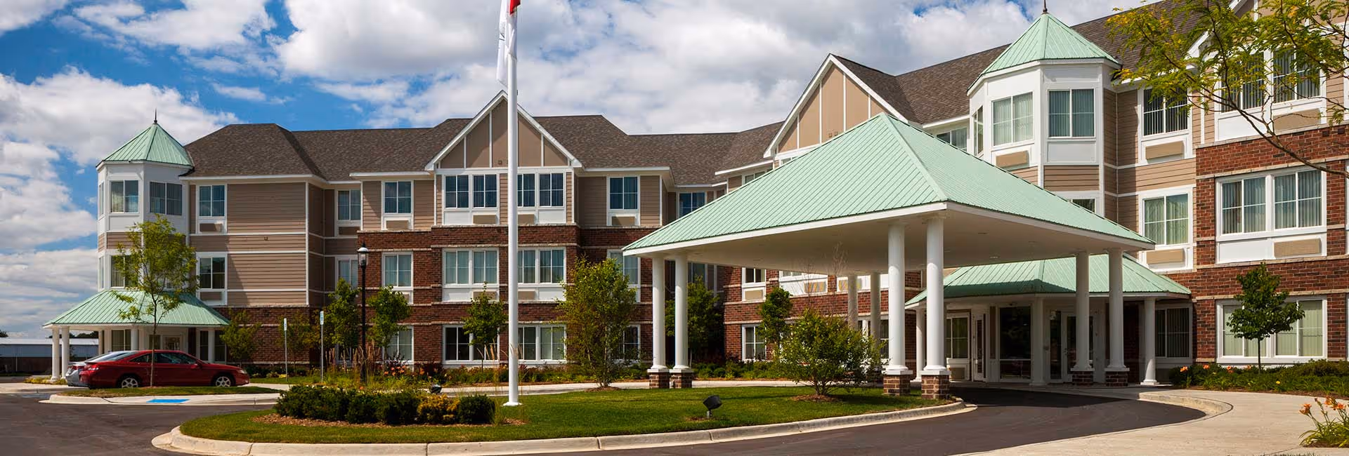 Exterior view of The Rivers Grosse Pointe senior living facility showing a large multi-story building with a covered entrance, green roofs, multiple windows, a flagpole, landscaped greenery, and a red car parked near the entrance under a partly cloudy sky.