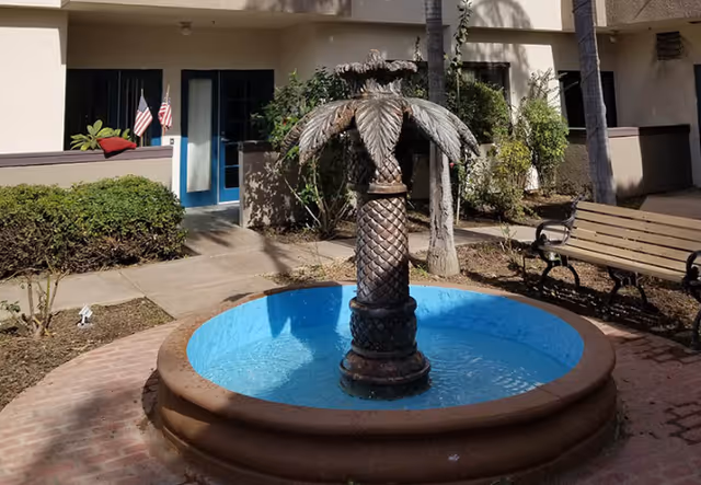 Outdoor courtyard area featuring a circular water fountain with a pineapple-shaped sculpture in the center. Surrounding the fountain are a brick pathway, a wooden bench with metal armrests, some bushes, and a building entrance with blue double doors and two small American flags on the left side.