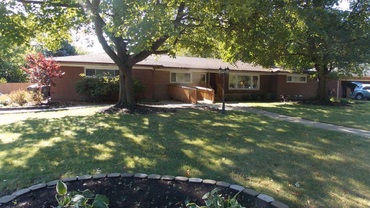 Single-story brick building with a sloped roof surrounded by green grass and large trees casting shadows. A paved walkway leads to a wooden ramp entrance. There is a lamp post near the walkway and a small garden bed with plants in the foreground.