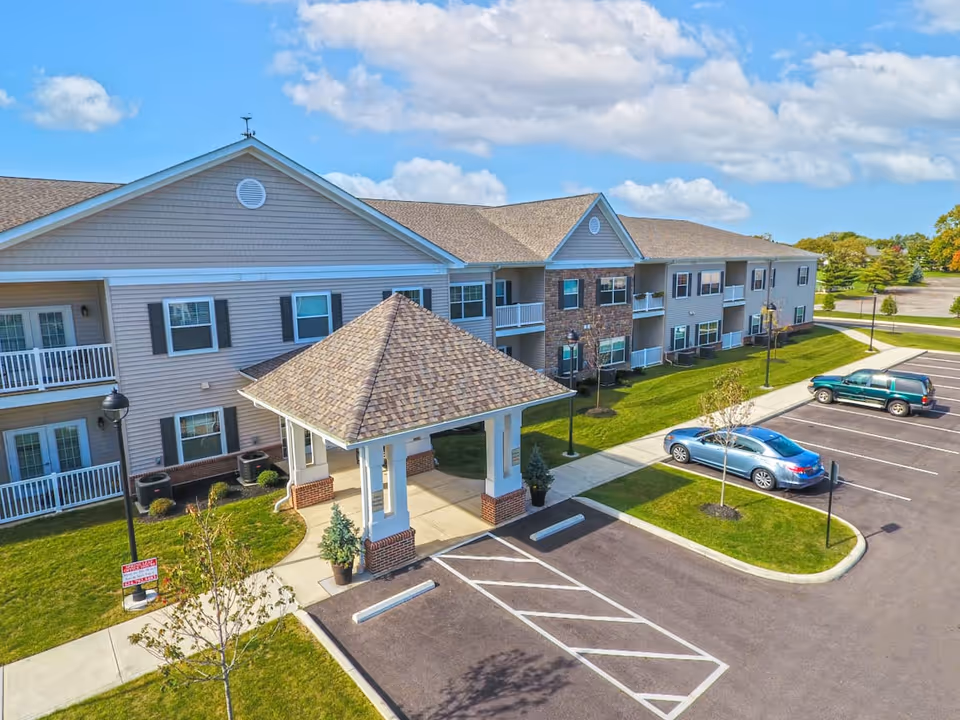 Exterior view of a two-story senior living facility building with beige siding and stone accents. The building has balconies and windows, a covered entrance with a peaked roof, a parking lot with a few cars, green lawns, and a partly cloudy blue sky.
