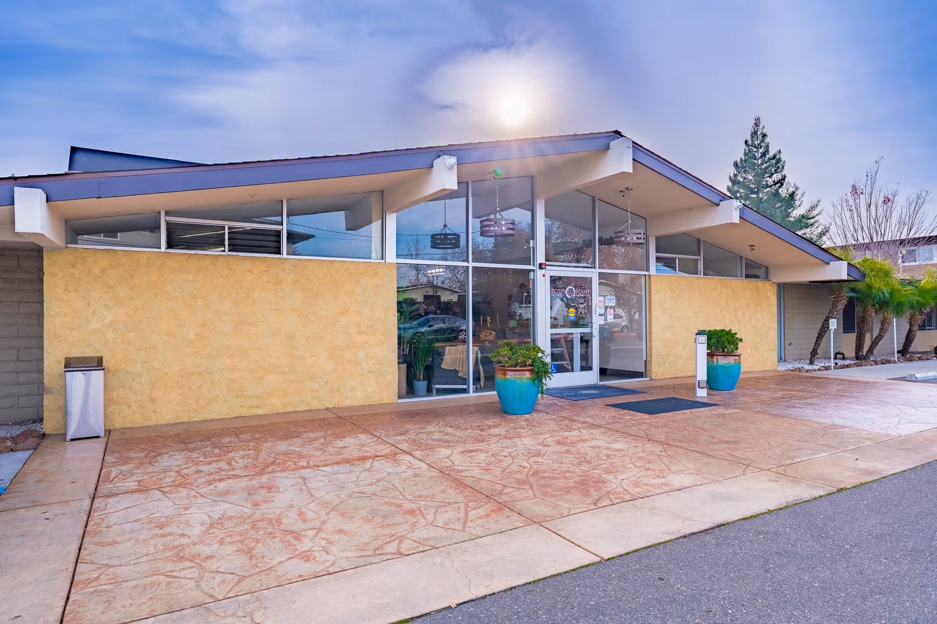 Exterior view of Roseville Point Health & Wellness Center showing a modern building entrance with large glass doors and windows, two large blue planters with green plants on either side of the entrance, a textured yellow wall, and a paved area in front under a partly cloudy sky with the sun visible.