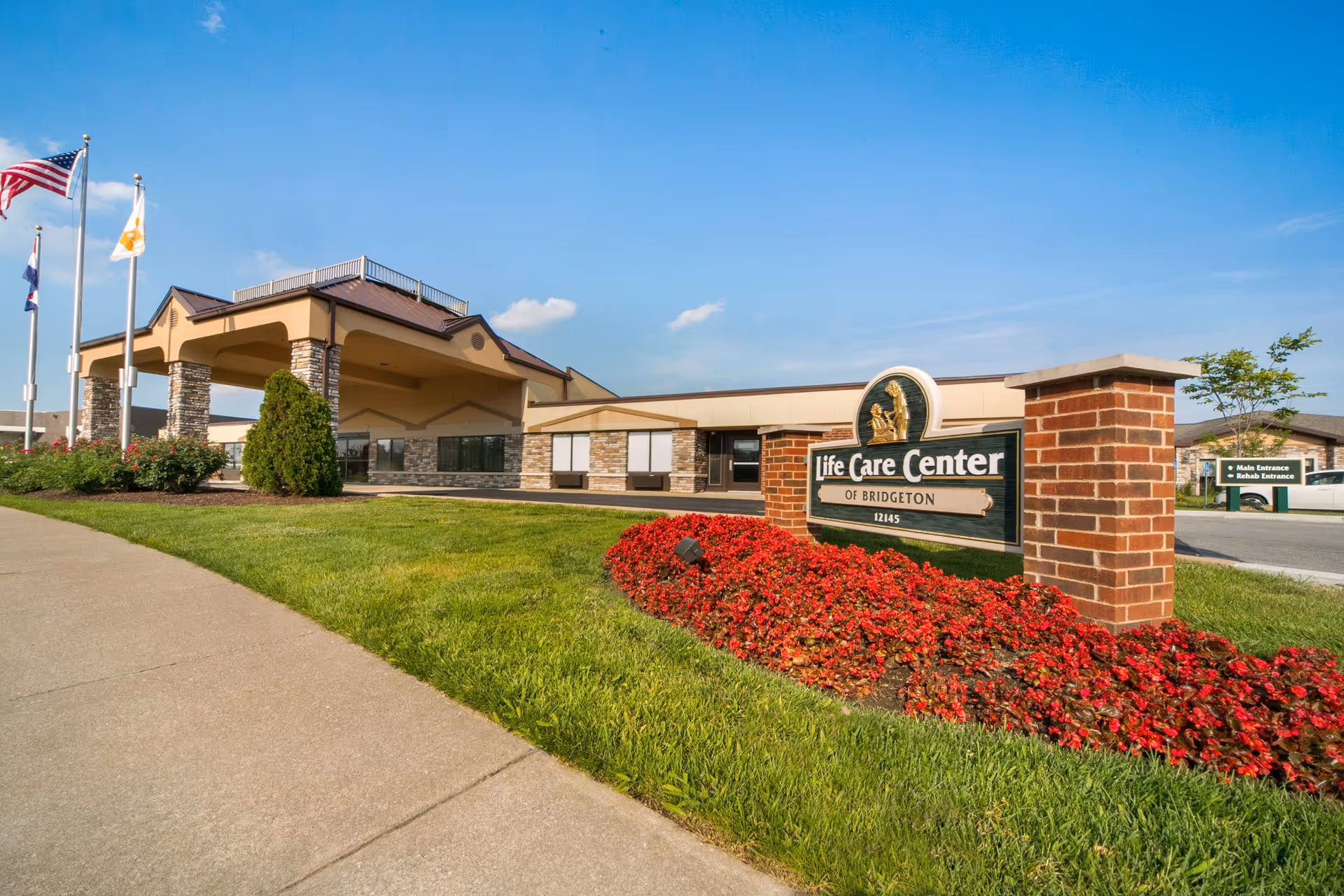 Exterior view of the Life Care Center of Bridgeton building with a brick sign in the foreground surrounded by red flowers and green grass. Three flagpoles with flags are visible on the left side, and the sky is clear and blue.