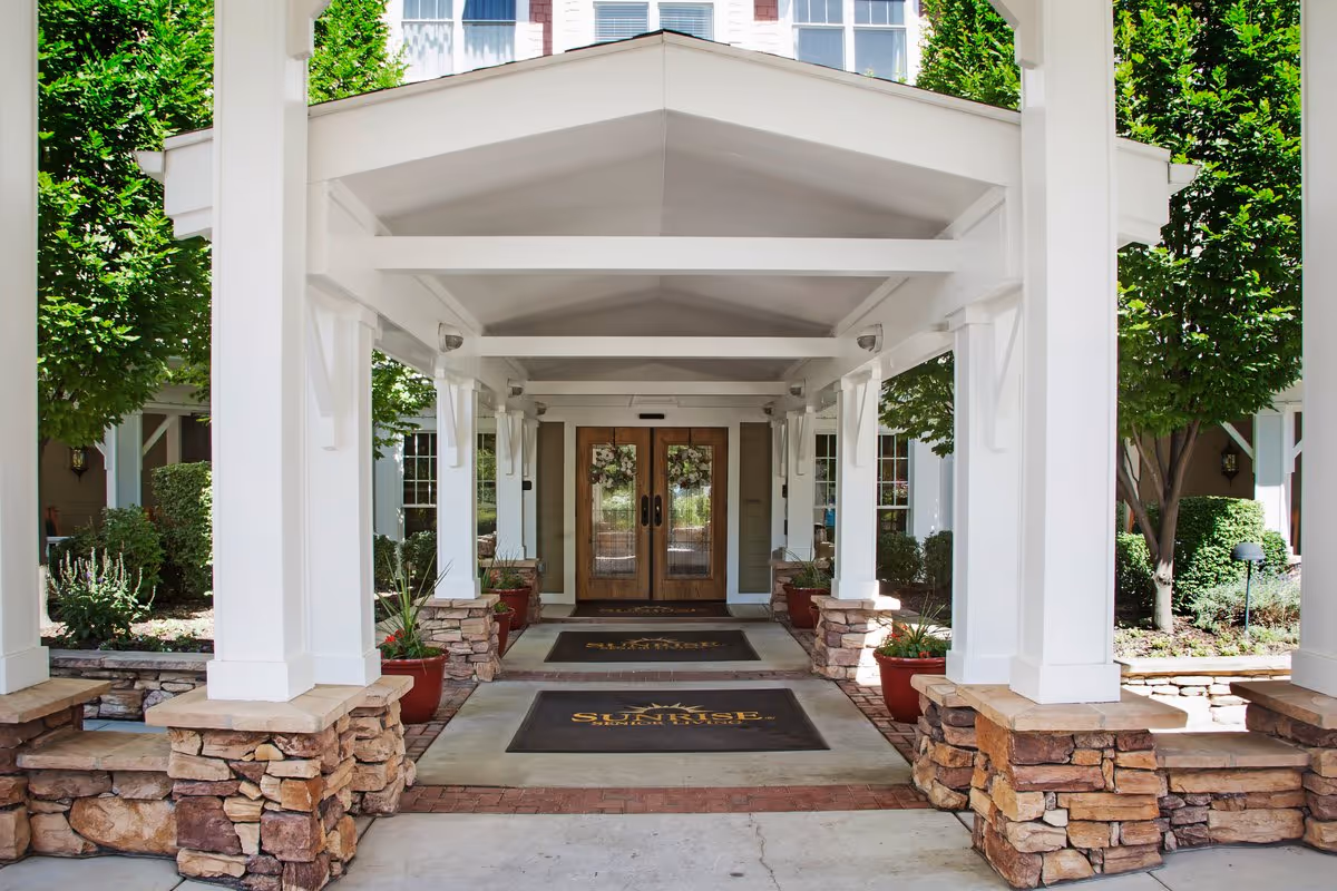 Covered entrance to a building with white pillars and stone bases, potted plants along the walkway, and double wooden doors decorated with wreaths. Two mats on the ground display the name 'Sunrise'.