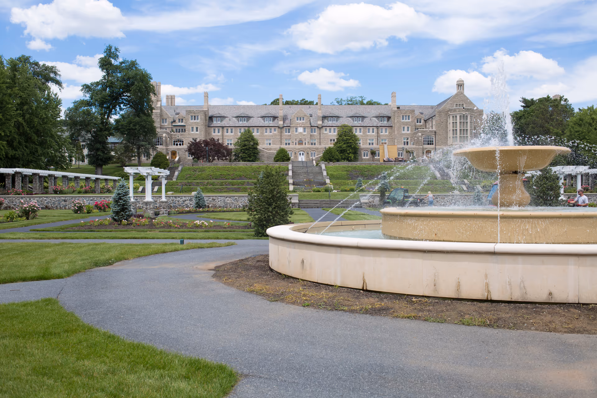 Large stone building with multiple windows and chimneys in the background, surrounded by well-maintained gardens and a large fountain in the foreground with water spraying. Paved pathways and green lawns are visible under a partly cloudy sky.