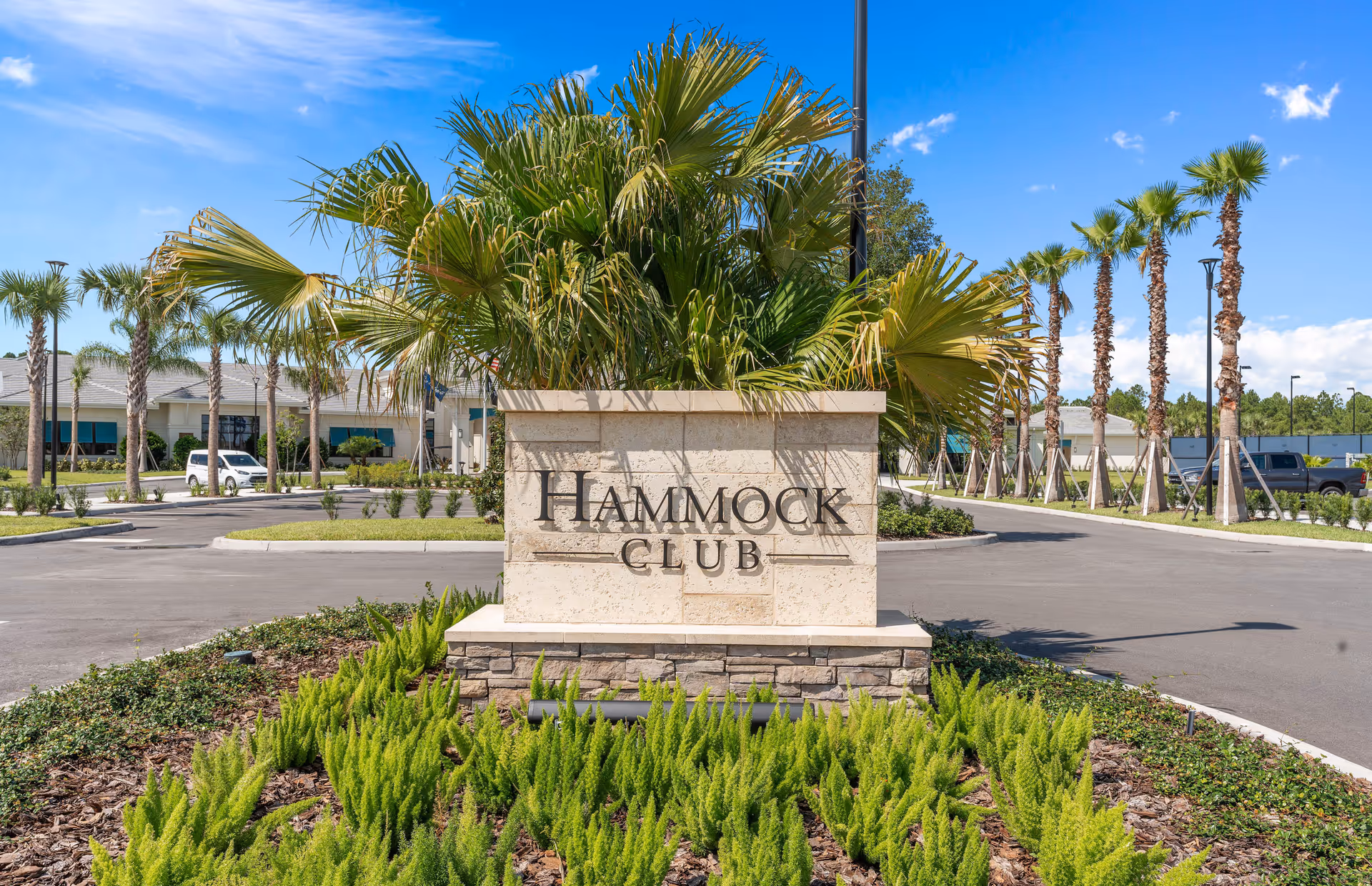 Stone sign with the text 'HAMMOCK CLUB' surrounded by green plants and palm trees, with a clear blue sky and buildings in the background.