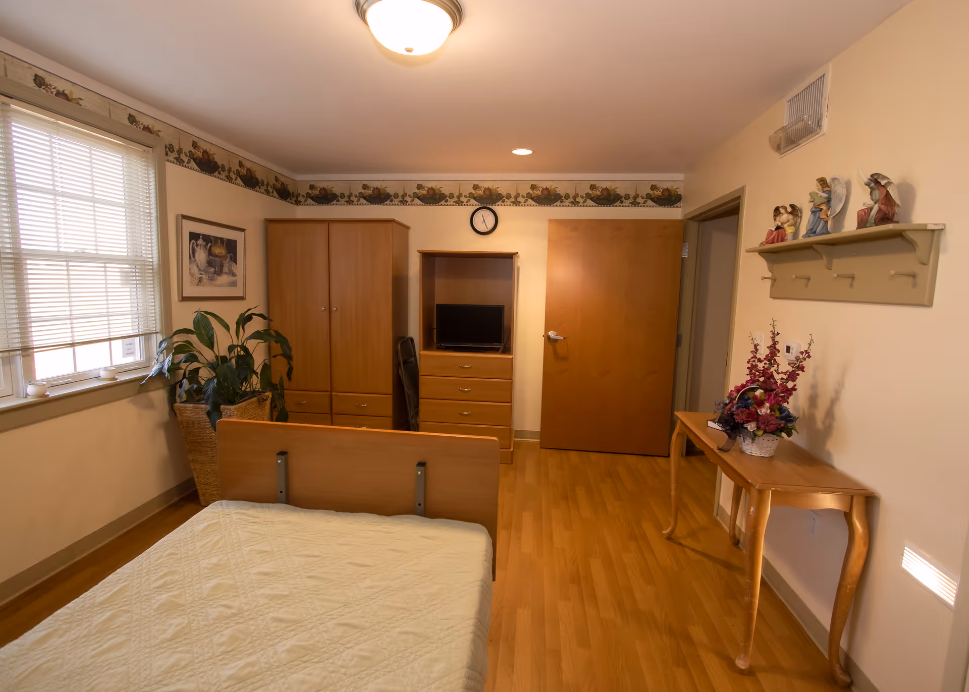 A simple, clean bedroom in a senior living facility with a bed covered in a white quilt, wooden furniture including a wardrobe, dresser with a small TV, and a table with a floral arrangement. There is a window with blinds, a plant in a wicker basket, a wall clock, and decorative figurines on a wall shelf.
