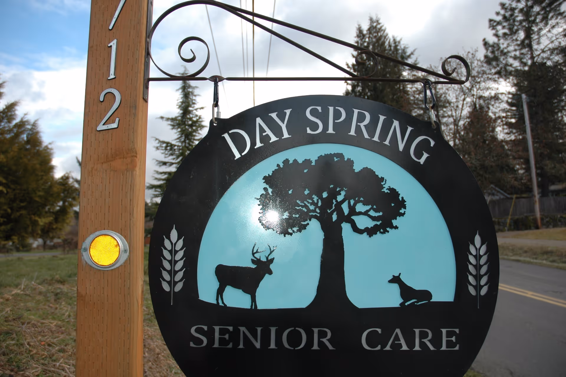 A hanging sign for Day Spring Senior Care attached to a wooden post with the number 712. The sign features silhouettes of a tree, a standing deer, and a resting deer against a light blue background with decorative wheat stalks on each side.