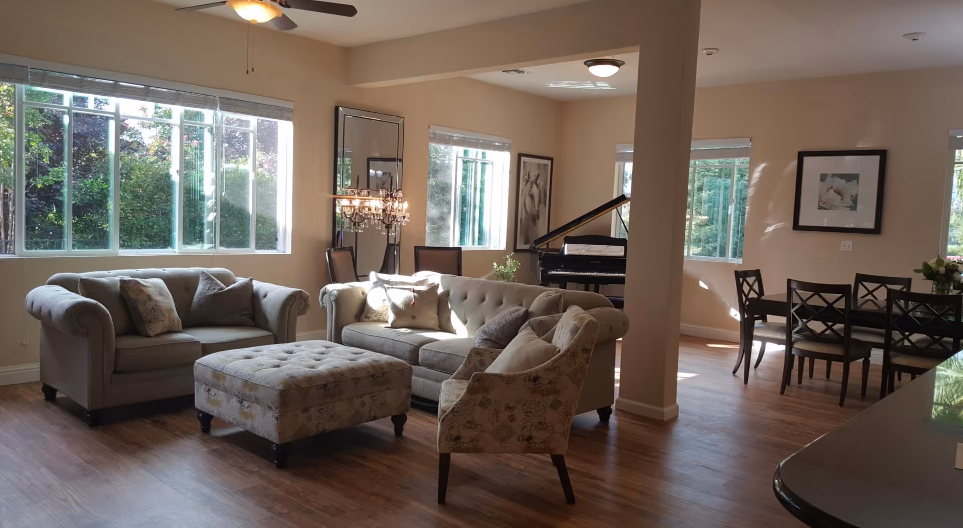 A bright and spacious living room area with large windows letting in natural light. The room features a beige sofa, a matching loveseat, an upholstered armchair, and a large ottoman in the center. There is a grand piano in the corner near the windows, a dining table with chairs to the right, and framed artwork on the walls. The floor is wooden, and the overall decor is warm and inviting.
