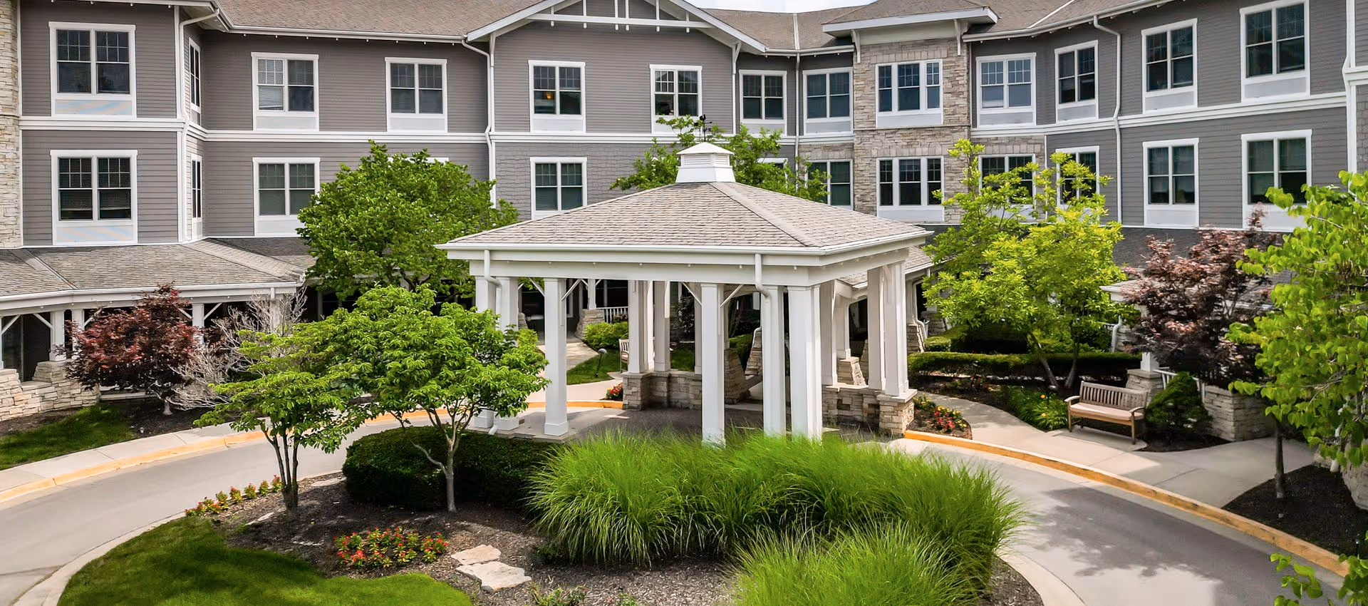 Exterior view of Hemsley House of Lenexa showing a large multi-story building with numerous windows, surrounded by landscaped greenery including trees, bushes, and flowers. In the foreground, there is a white gazebo structure with a shingled roof, situated in the middle of a circular driveway.
