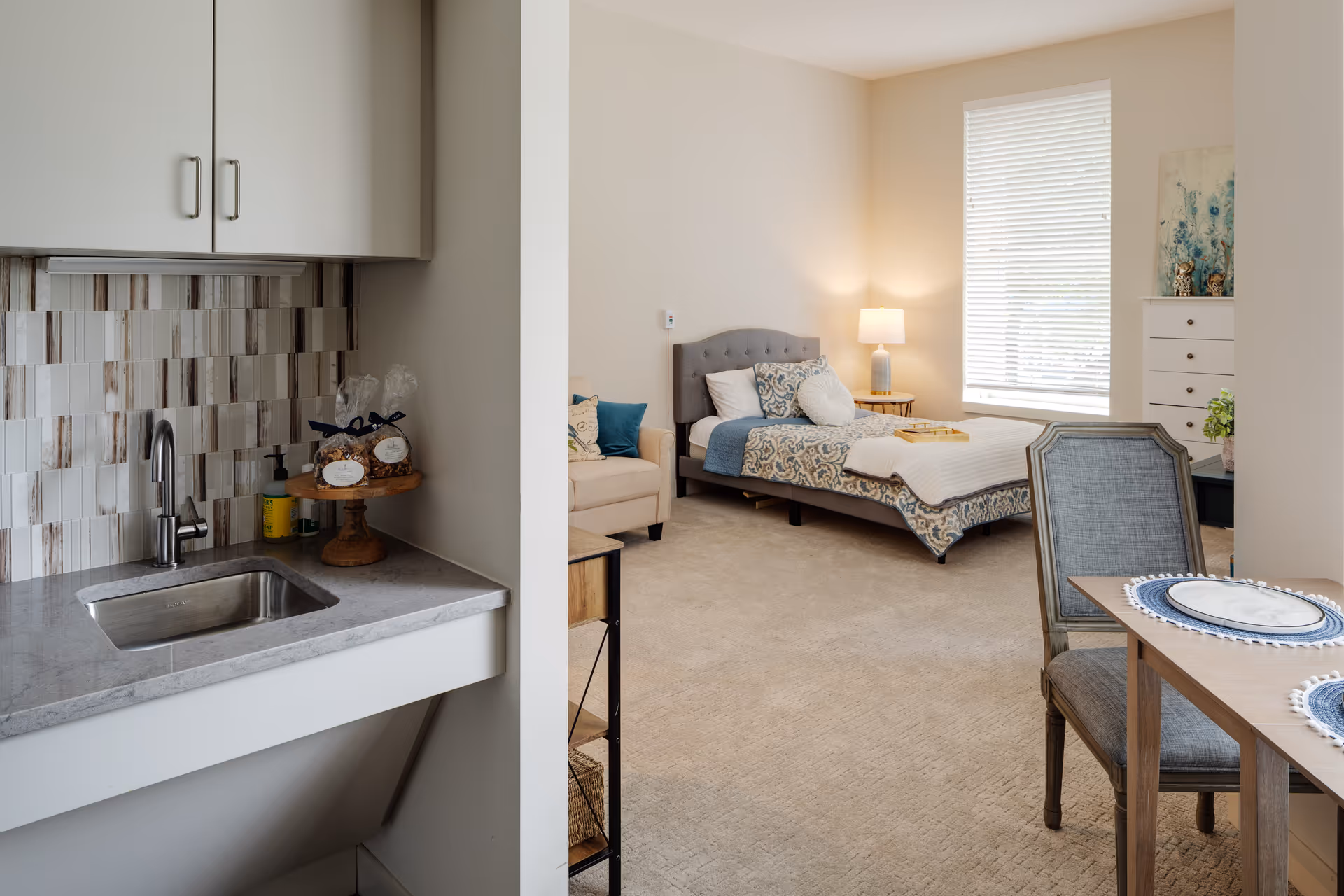 A cozy senior living studio apartment featuring a small kitchenette with a sink and tiled backsplash on the left, a neatly made bed with patterned bedding and pillows in the center, a beige armchair, a bedside table with a lamp, a window with blinds, a white dresser with decorative items, and a dining table with two chairs and place settings on the right.