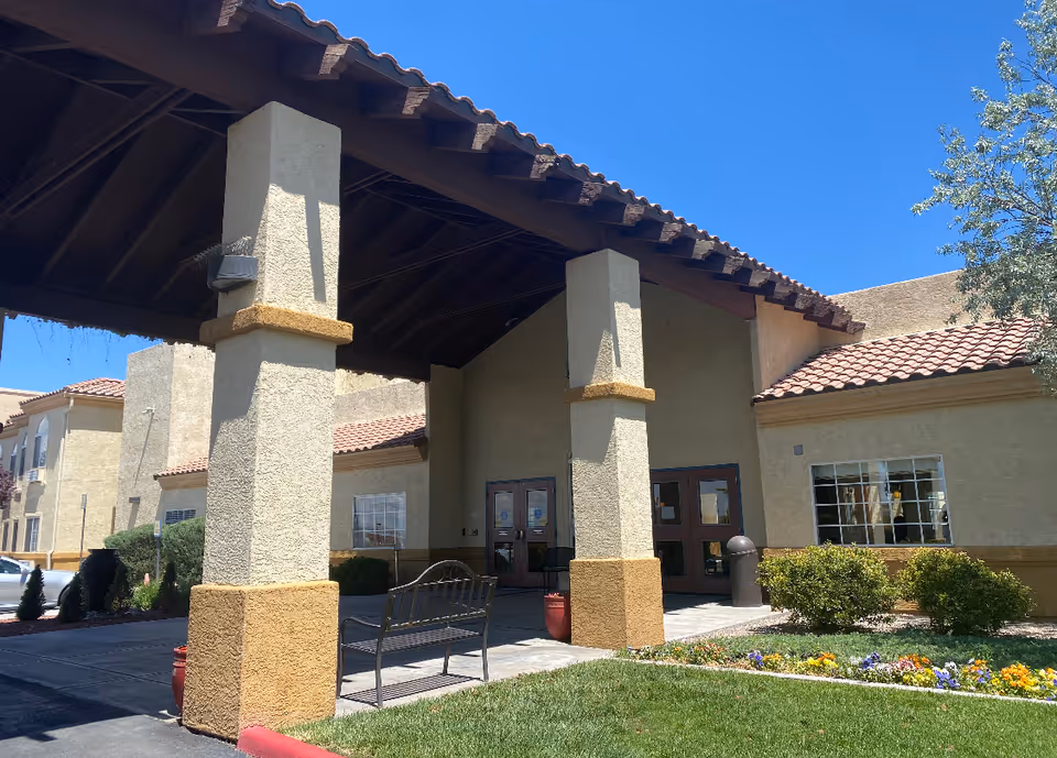 Exterior view of the entrance to a senior living facility with a covered driveway supported by large beige columns, a bench, landscaped bushes, and colorful flowers under a clear blue sky.