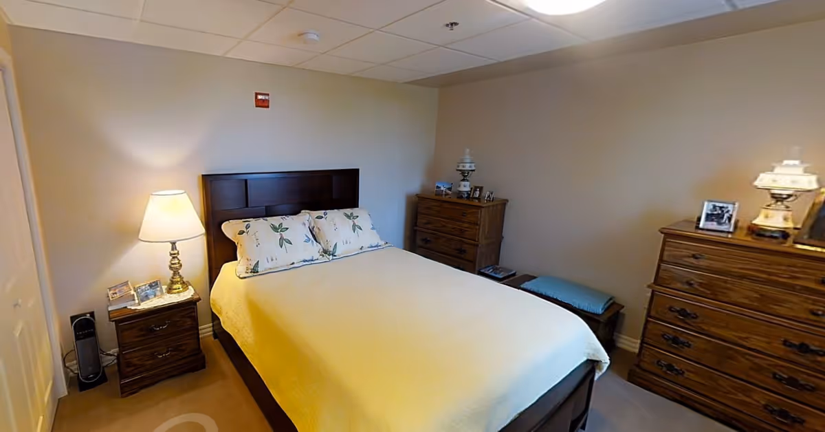A cozy bedroom with a double bed covered in a light yellow bedspread and two floral pillows. There are two wooden dressers, one on each side of the bed, each with a lamp and framed photos on top. A small bench with a blue cushion is placed against the wall near one dresser. The walls are painted beige and the room has a soft, warm lighting.