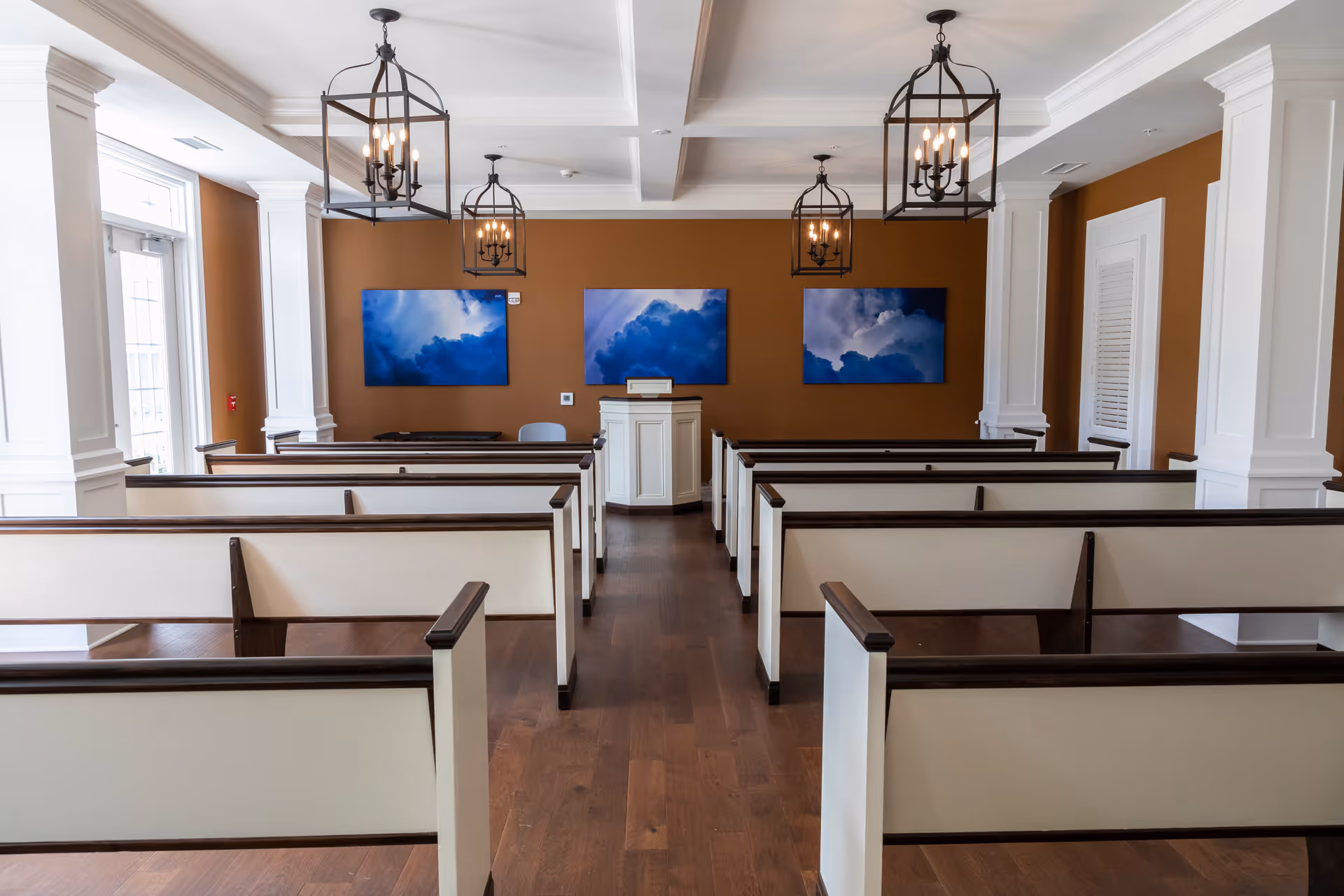Interior view of a room with white wooden pews arranged in rows facing a small podium against a brown wall. The room has wooden flooring, white columns, and three blue cloud-themed paintings on the wall behind the podium. Four black lantern-style chandeliers hang from the ceiling.