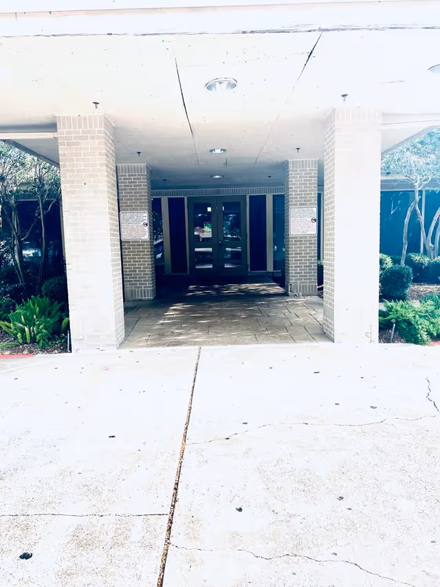 Entrance area of a building with a covered driveway supported by brick pillars, leading to double glass doors. There are small bushes and trees on either side of the entrance.