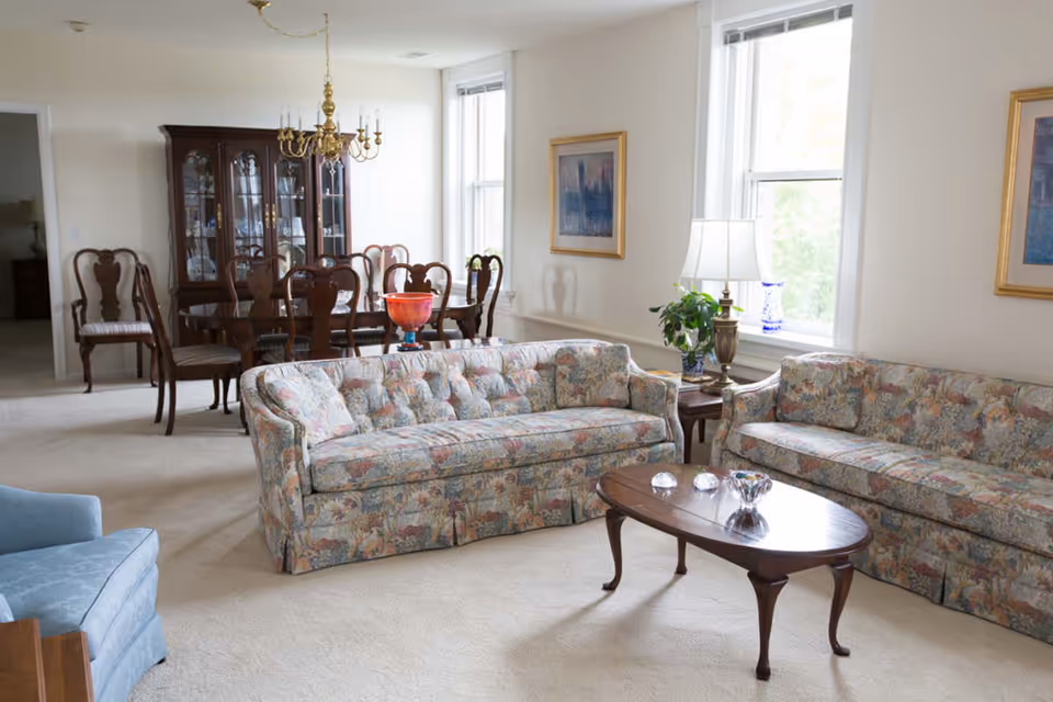 A bright living room with two floral patterned sofas, a wooden coffee table with decorative items, a side table with a lamp and plant, and a dining area in the background featuring a wooden dining table with chairs and a china cabinet.