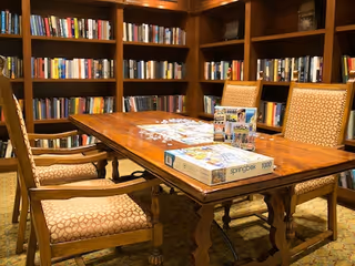 A cozy library room with wooden bookshelves filled with books lining the walls. In the center, there is a wooden table with a puzzle box and puzzle pieces on it, surrounded by four upholstered chairs with patterned fabric.